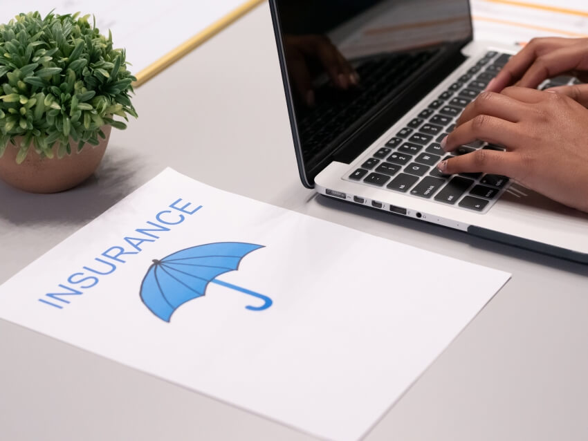 Man Typing in a Laptop with Printred Insurance and Umbrella on Paper on its Side