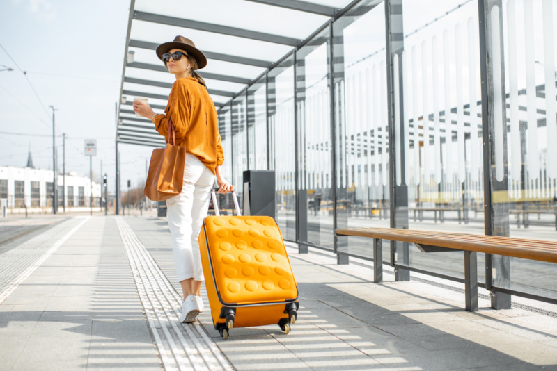 Young female traveler walking with a yellow suitcase at the modern transport stop outdoors, back view.