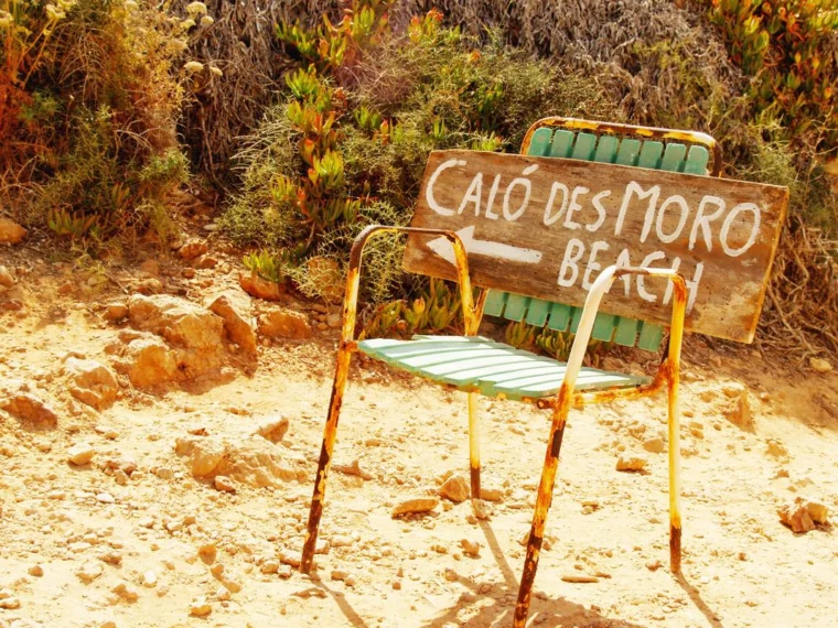 Rustic sign for Caló des Moro Beach on a weathered chair amidst rocky terrain and lush greenery.