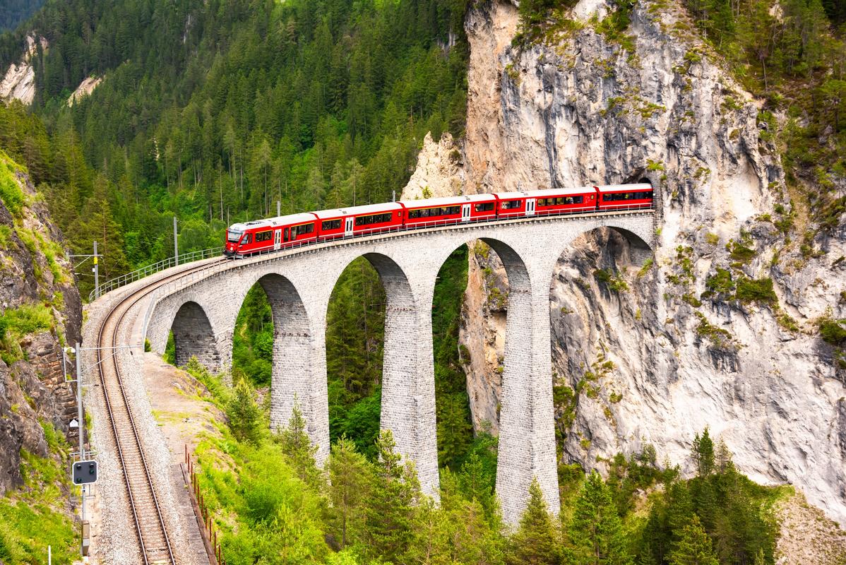 Red Bernina Railway train crossing viaduct in lush Swiss Alps forest.