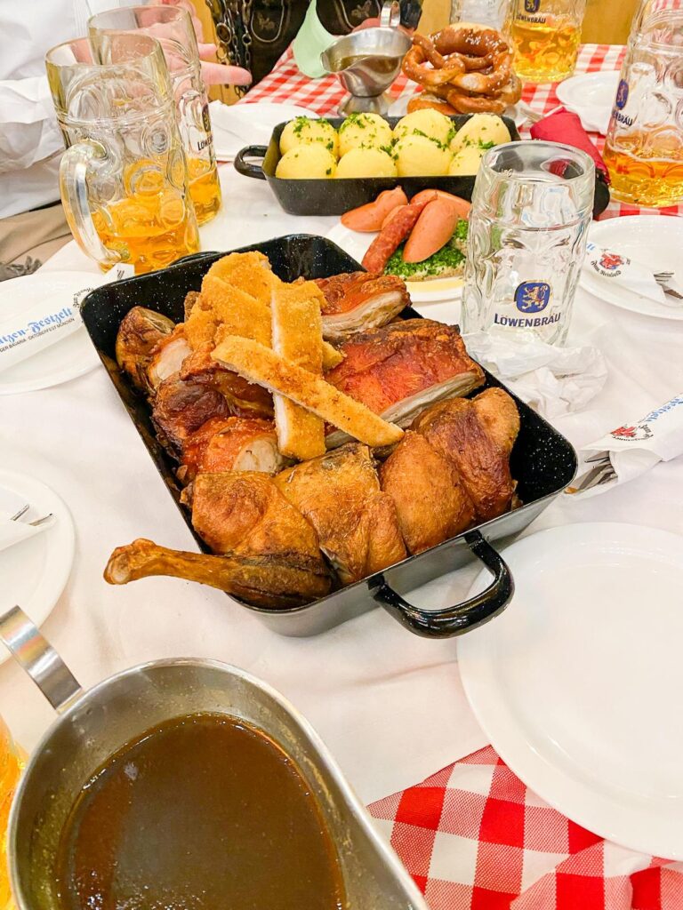 Authentic German feast at Oktoberfest in Munich with Schweinshaxe, potato dumplings, sausages, pretzel, and Löwenbräu beer on a checkered table.