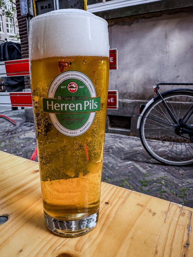 Bamberg Herren Pils beer on wooden table in Amsterdam, showcasing German brewing excellence and European cultural flair.