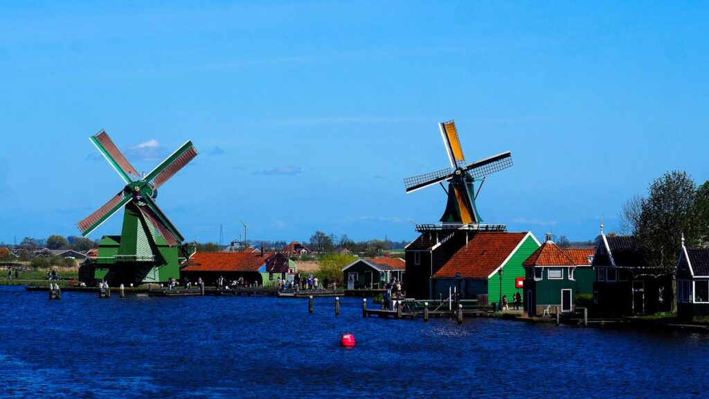 Historic green and yellow windmills of Zaanse Schans by a river with traditional Dutch wooden houses and a clear blue sky in the background.