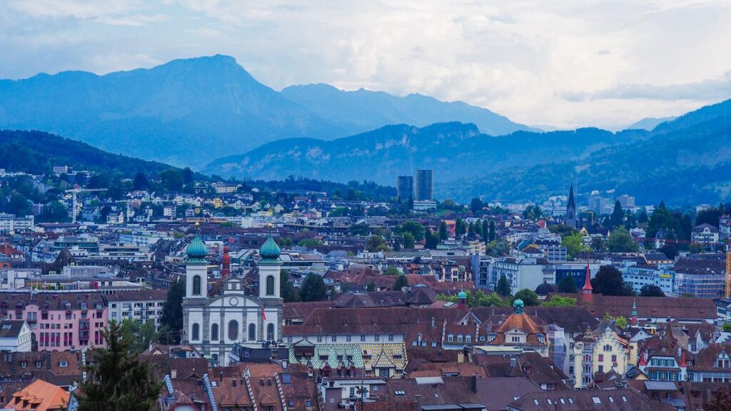 Panoramic view of Lucerne cityscape with Alpine backdrop, showcasing historical and modern architecture, Swiss rooftops, church with green domes, and misty Alps.