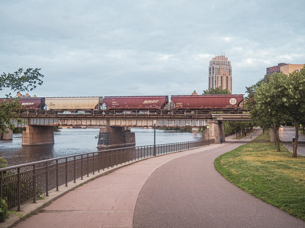 Alt text: Scenic evening stroll along Mississippi River, Minneapolis skyline & BNSF freight train.