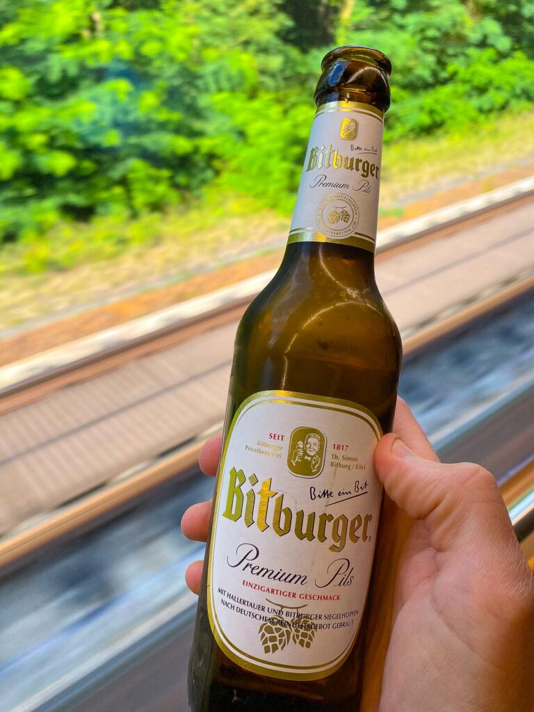 Traveler enjoying Bitburger Premium Pils beer on scenic train journey through German landscape.