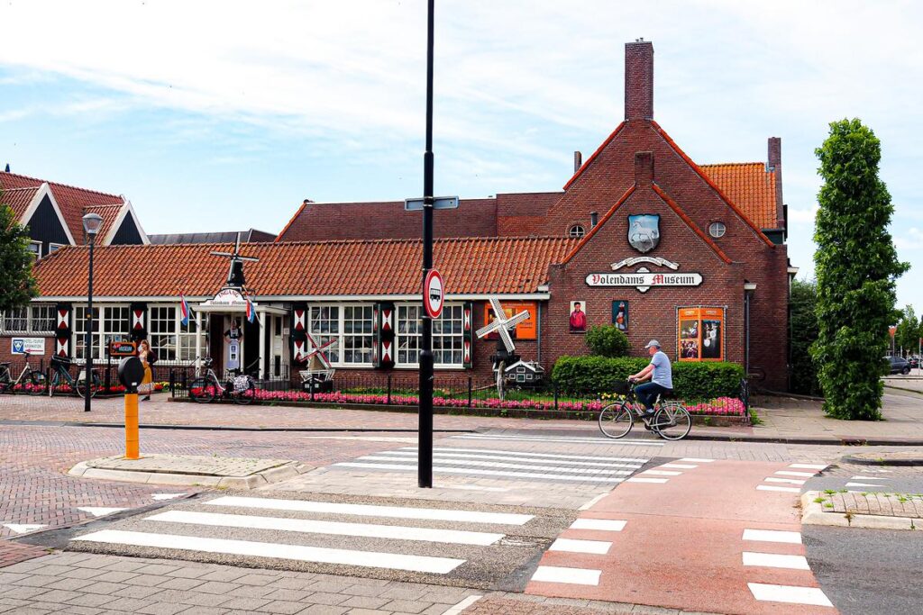 A bicyclist rides by the traditional Dutch architecture of the Volendams Museum decorated with vibrant flowers, under a sunny sky, reflecting the culture and lifestyle of Volendam, The Netherlands.